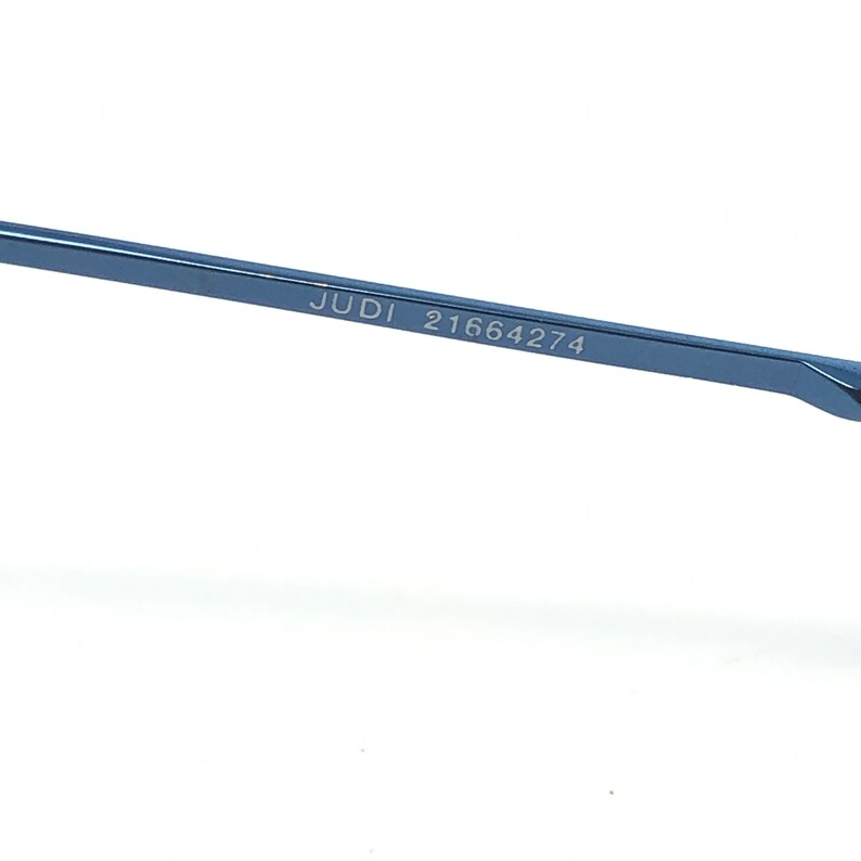 May include: Close-up of a section of blue eyeglasses. The text "JUDI 21664274" is printed on the frame. The glasses are set against a white background.