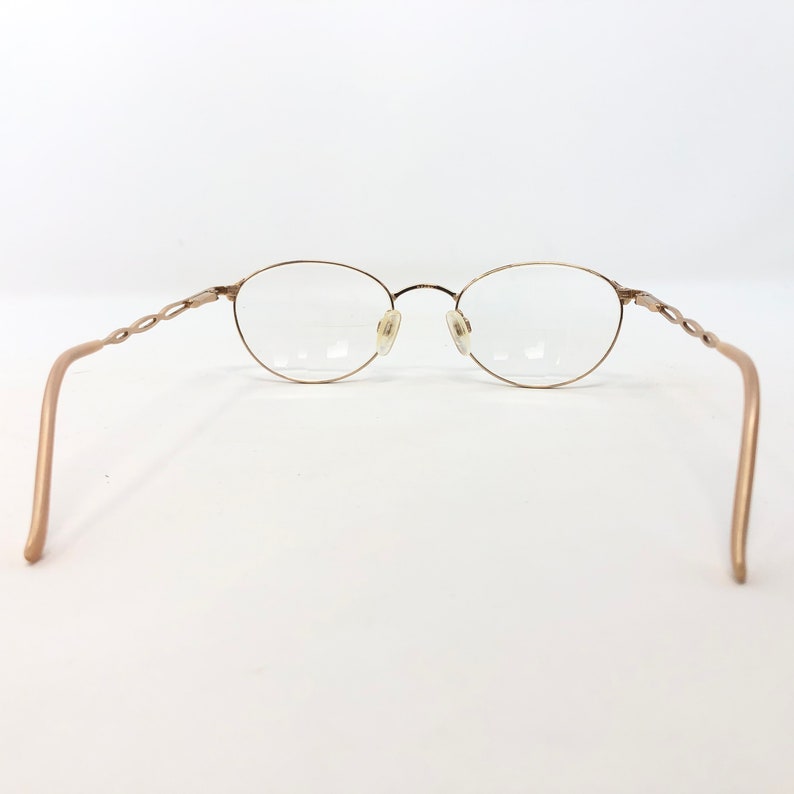 K&ouml;nnte beinhalten: Goldfarbene Brille mit ovalen Gl&auml;sern und einem filigranen, gedrehten Design an den B&uuml;geln. Die Fassungen haben einen dezenten metallischen Glanz, und die Gl&auml;ser sind klar. Das Gesamtdesign ist minimalistisch und elegant.