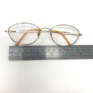 Puede incluir: Gafas ovaladas con montura de metal plateado y lentes transparentes. Las patillas son de color marr&oacute;n claro. Una regla se coloca junto a las gafas, mostrando medidas en cent&iacute;metros.