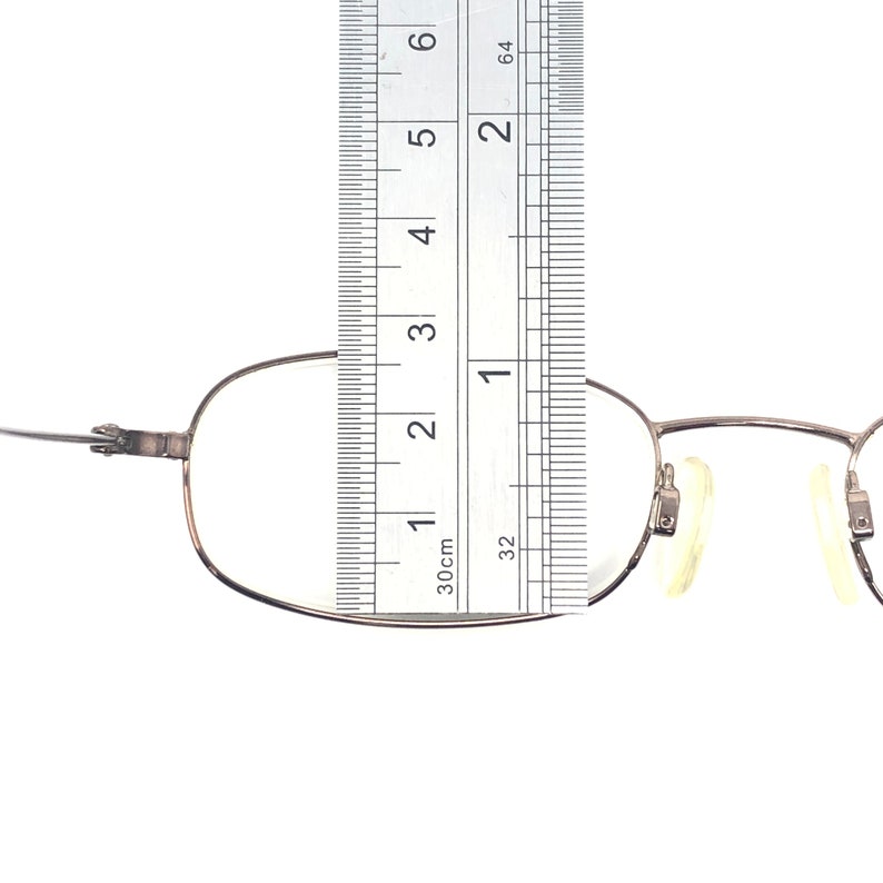 Puede incluir: Gafas plateadas con montura rectangular. Una regla se coloca sobre las gafas, mostrando medidas en cent&iacute;metros. Las gafas tienen almohadillas nasales y un dise&ntilde;o de bisagra.