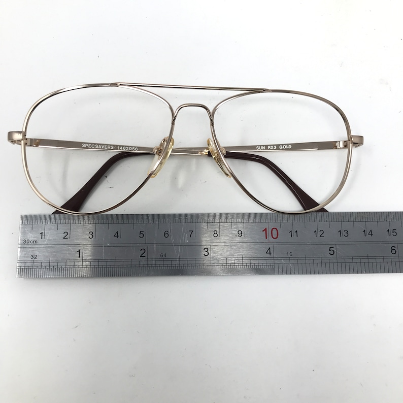 May include: Gold-tone aviator eyeglasses with clear lenses. The glasses have a double bridge and brown earpieces. The text "SPECSAVERS 1462056" and "SUN RX3 GOLD" are visible on the frame. A ruler shows the glasses are approximately 5.5 inches wide.