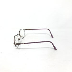 Puede incluir: Un par de gafas con lentes ovaladas y una montura met&aacute;lica fina y oscura. Las patillas son de color morado liso. Las gafas est&aacute;n sobre un fondo blanco liso.