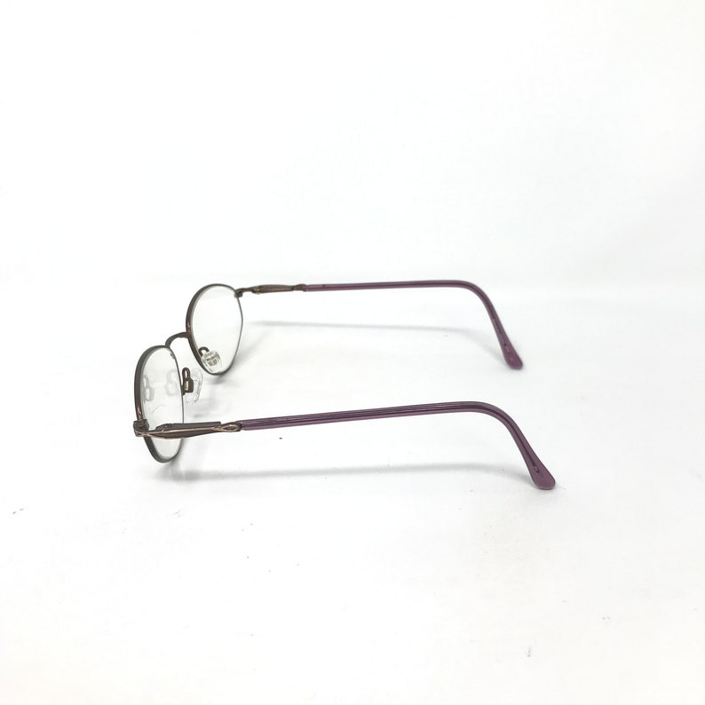 Puede incluir: Un par de gafas con lentes ovaladas y una montura met&aacute;lica fina y oscura. Las patillas son de color morado liso. Las gafas est&aacute;n sobre un fondo blanco liso.
