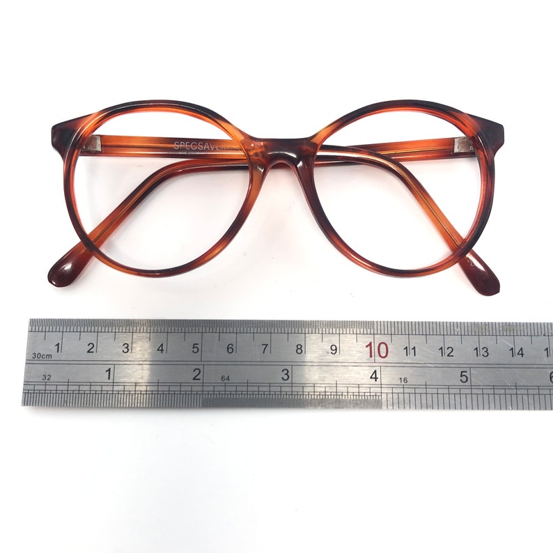 May include: Brown tortoiseshell eyeglasses with round frames. The glasses are placed on a white surface next to a ruler. The text "SPECSAVERS" is visible on the frame. The ruler measures up to 15 inches.