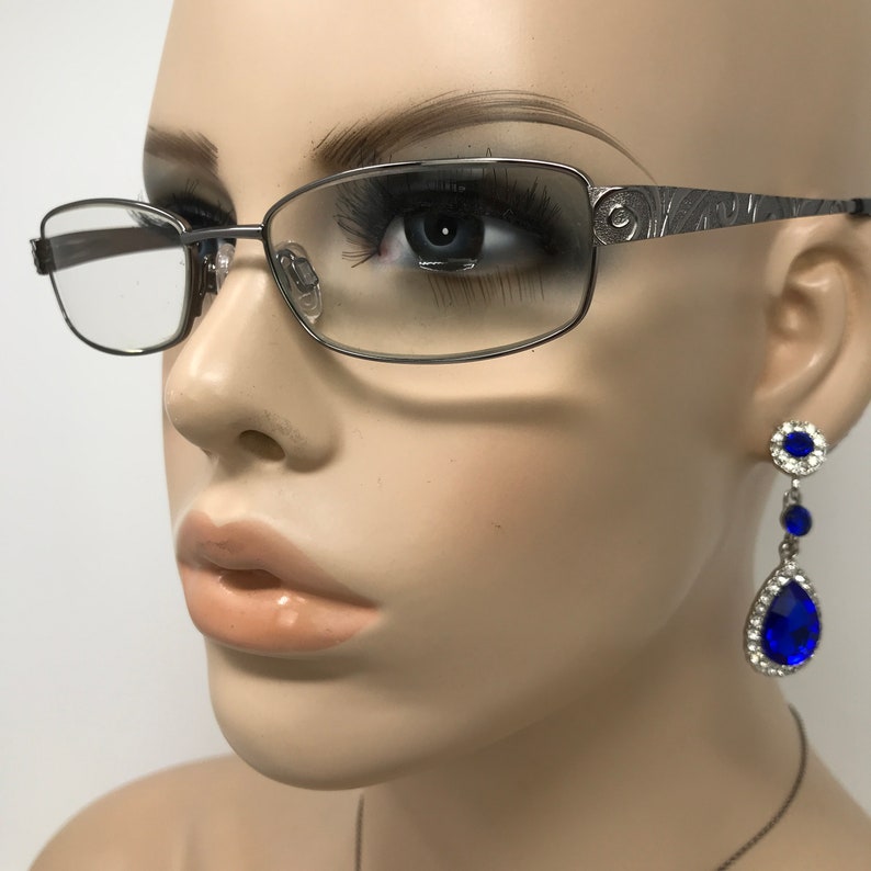 May include: Silver-framed eyeglasses with rectangular lenses and decorative temples. The image also shows a pair of dangling earrings with blue teardrop-shaped gems and smaller blue stones. The jewelry is set in silver.