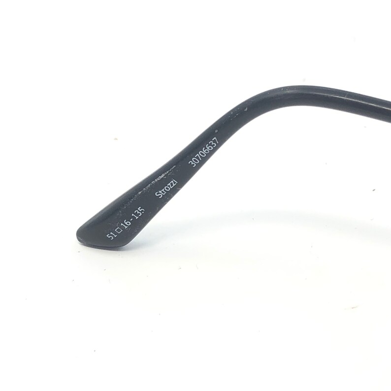 May include: Close-up of a black eyeglass arm with the text "Strozzi", "30706637", and measurements "51 16-135". The arm is curved and made of a matte material. The background is white.