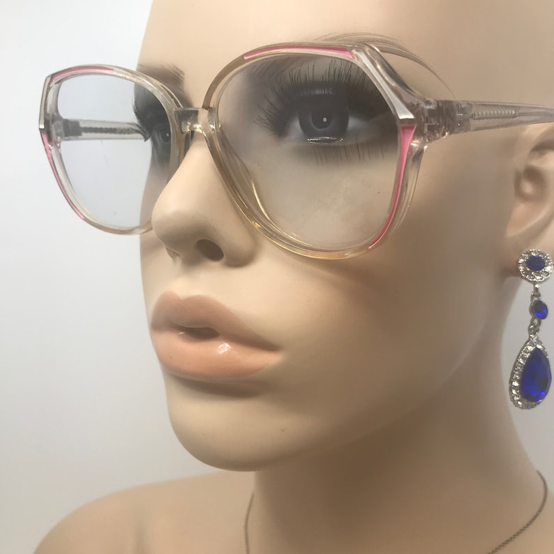 May include: Clear-framed eyeglasses with pink accents. The glasses have a large, square shape and are displayed on a mannequin. A pair of blue and silver drop earrings are also visible.
