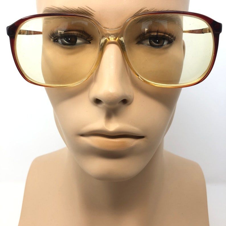 May include: Vintage eyeglasses with a clear and brown frame and yellow-tinted lenses. The glasses have a rectangular shape and are displayed on a mannequin head. The frames have a gradient color, transitioning from brown to clear.