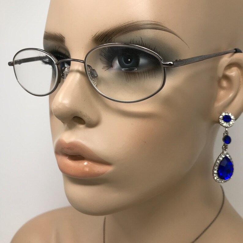 Op de afbeelding: Een mannequin draagt een ovale bril met een zilveren montuur. De mannequin draagt ook oorhangers met blauwe, druppelvormige edelstenen en kleinere blauwe edelstenen omringd door heldere stenen.