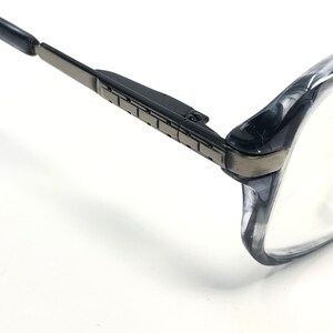 May include: Close-up of spectacles with a black and grey frame. The glasses have a rectangular shape with a dark grey metal hinge and a clear lens. The frame is a translucent grey colour.
