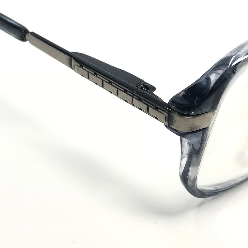 May include: Close-up of spectacles with a black and grey frame. The glasses have a rectangular shape with a dark grey metal hinge and a clear lens. The frame is a translucent grey colour.