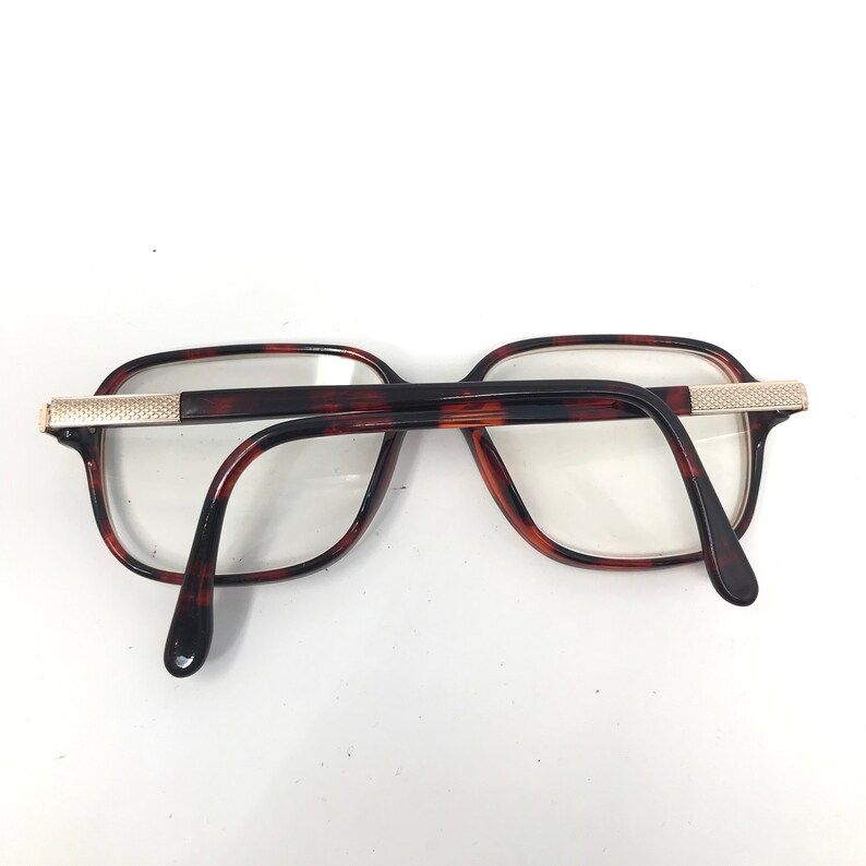 May include: Eyeglasses with a tortoiseshell pattern frame and clear lenses. The rectangular frames have a gold-colored metal accent on the temples. The arms are dark brown and curve down to the ear pieces.