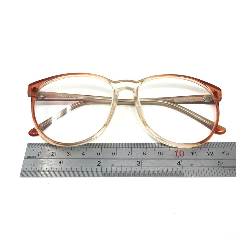 May include: Clear eyeglasses with a gradient brown frame. The glasses have a round shape and are measured with a ruler. The text "SPECSAVERS" is visible on the frame. The ruler shows a length of approximately 5 inches.