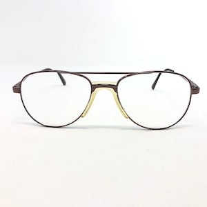 May include: A pair of aviator-style eyeglasses with a dark brown metal frame. The lenses are clear, and the nose pads are a light yellow color. The glasses have a double bridge and thin temples.