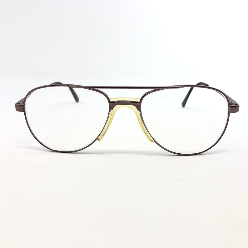 May include: A pair of aviator-style eyeglasses with a dark brown metal frame. The lenses are clear, and the nose pads are a light yellow color. The glasses have a double bridge and thin temples.