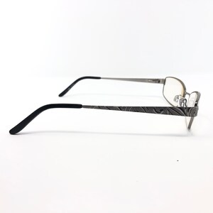 May include: Silver-toned eyeglasses with a decorative floral pattern on the temples. The frames have a rectangular shape with clear lenses and black earpieces. The glasses are set against a white background.
