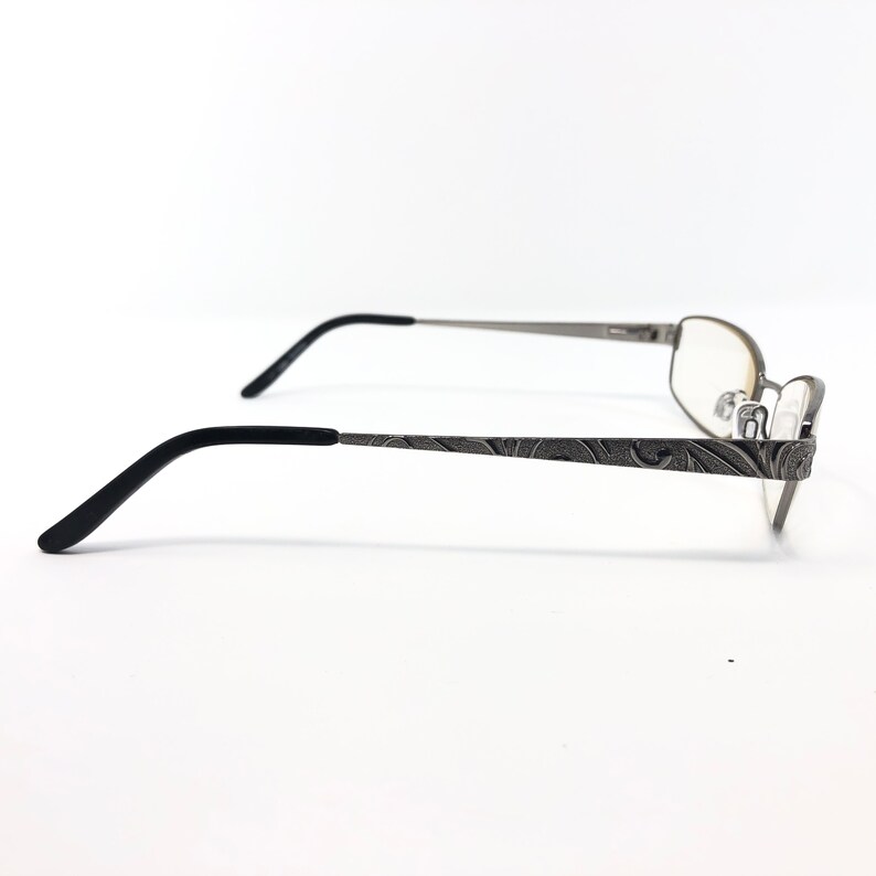May include: Silver-toned eyeglasses with a decorative floral pattern on the temples. The frames have a rectangular shape with clear lenses and black earpieces. The glasses are set against a white background.