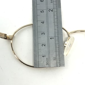 Pu&ograve; includere: Primo piano di una montatura di occhiali color oro con un righello che misura il diametro della montatura. La montatura &egrave; rotonda e ha i naselli. Il righello mostra le misure in centimetri.