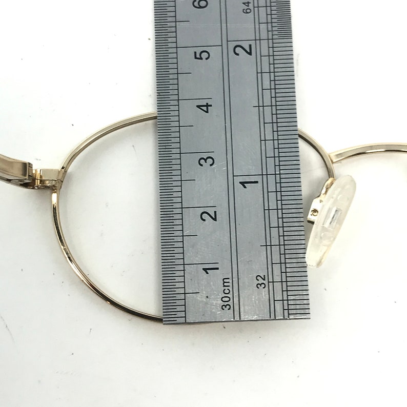 Pu&ograve; includere: Primo piano di una montatura di occhiali color oro con un righello che misura il diametro della montatura. La montatura &egrave; rotonda e ha i naselli. Il righello mostra le misure in centimetri.