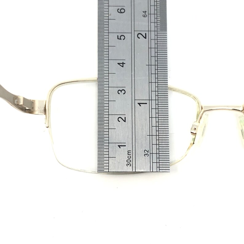 Pu&ograve; includere: Occhiali rettangolari color argento con un righello che mostra una misura di circa 2,5 cm. Gli occhiali hanno una montatura sottile e sono su uno sfondo bianco. Il righello &egrave; contrassegnato in pollici e centimetri.