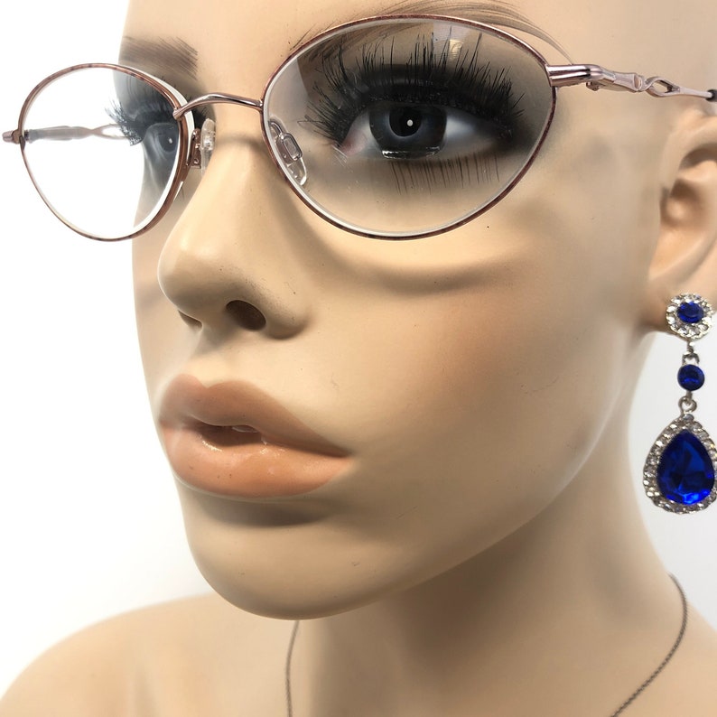 Puede incluir: Maniqu&iacute; con gafas ovaladas de oro rosa con lentes transparentes. El maniqu&iacute; tambi&eacute;n lleva un par de pendientes colgantes con una piedra azul en forma de l&aacute;grima y piedras azules y transparentes m&aacute;s peque&ntilde;as.