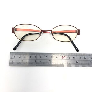 May include: Oval-shaped eyeglasses with a maroon frame and black temples. The lenses are clear, and a ruler is placed next to the glasses for scale. The text "VOS MO HARMONY 784438" is printed on the temple.