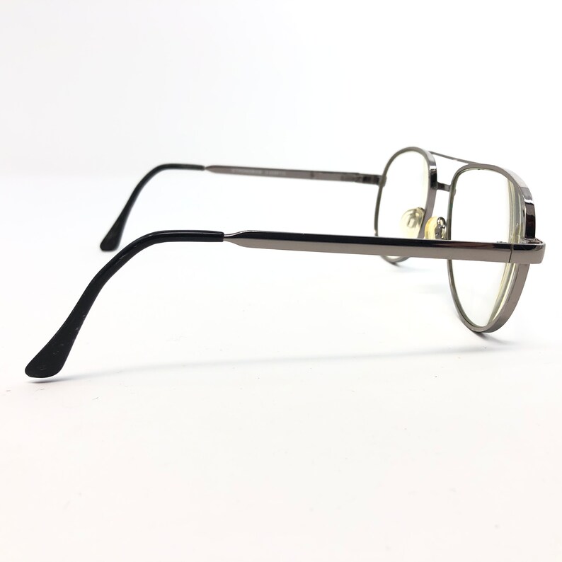 Peut inclure: Lunettes de vue aviateur de couleur canon de fusil avec des verres transparents. Les montures ont un double pont et des branches fines. Les branches sont noires. Les lunettes sont sur fond blanc.