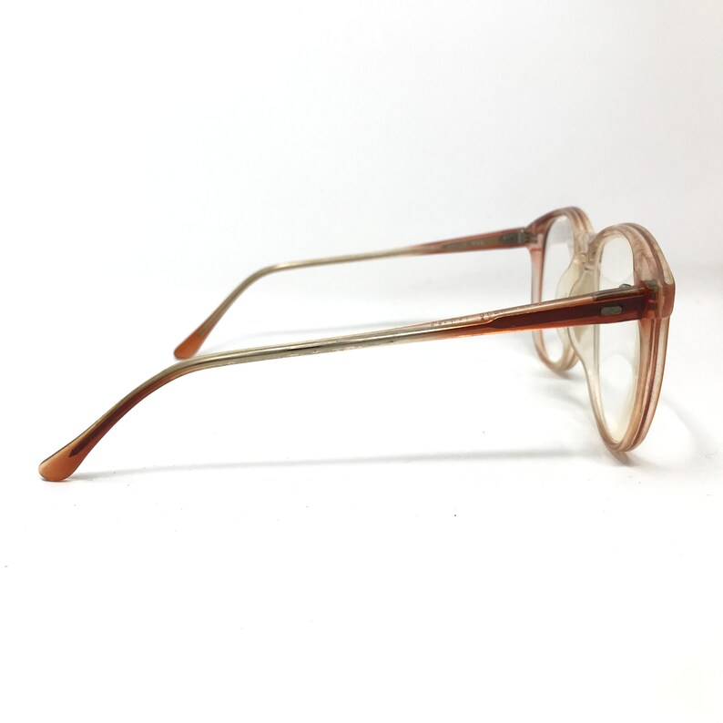 May include: A pair of vintage eyeglasses with a light brown frame and clear lenses. The glasses have a rectangular shape with a slightly rounded edge. The temples are a gradient of light brown to silver.