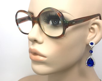May include: Round-framed spectacles with a tortoiseshell pattern. The glasses are worn by a mannequin. Dangling earrings with blue teardrop-shaped gems and silver accents are also visible.