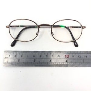 Puede incluir: Gafas ovaladas con una fina montura met&aacute;lica de color bronce. Las lentes son transparentes y las patillas son negras. Una regla met&aacute;lica con cent&iacute;metros se coloca debajo.