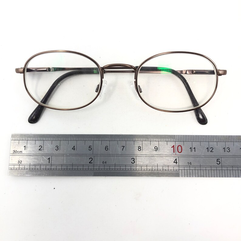 Puede incluir: Gafas ovaladas con una fina montura met&aacute;lica de color bronce. Las lentes son transparentes y las patillas son negras. Una regla met&aacute;lica con cent&iacute;metros se coloca debajo.