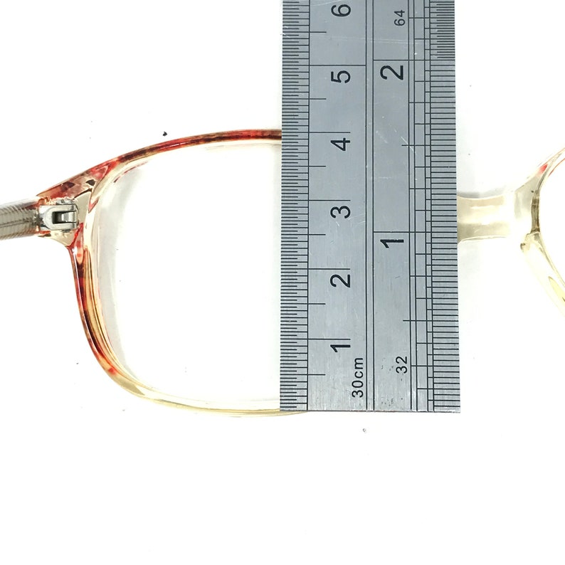 May include: Close-up of eyeglasses with a clear frame and a red and brown patterned accent. The frame measures approximately 2 inches in length. The glasses are positioned next to a ruler for measurement.