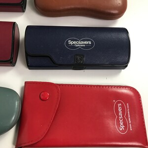 May include: Assortment of eyeglass cases in various colors. A navy blue case features the "Specsavers Opticians" logo. A red case has a button closure and the "Specsavers" logo. Other cases are burgundy, brown, and teal.