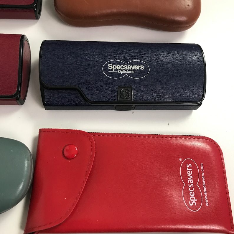 May include: Assortment of eyeglass cases in various colors. A navy blue case features the "Specsavers Opticians" logo. A red case has a button closure and the "Specsavers" logo. Other cases are burgundy, brown, and teal.