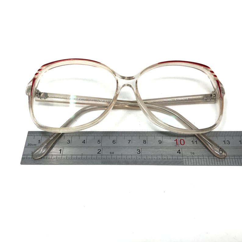 May include: Clear-framed eyeglasses with a rectangular shape and red accents on the upper frame. The glasses are measured with a ruler, showing a length of approximately 5 inches.