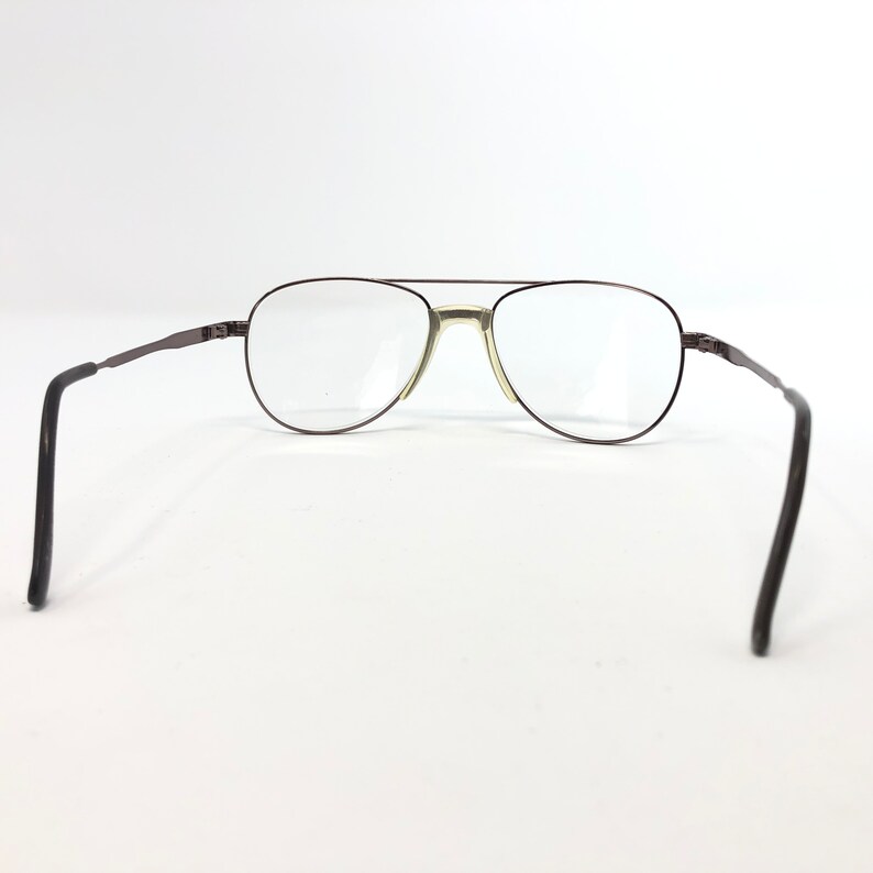 May include: Aviator-style eyeglasses with a dark brown metal frame and clear lenses. The glasses have a double bridge and thin temples that curve at the ends. The lenses are oval-shaped and the frame is a dark brown color.