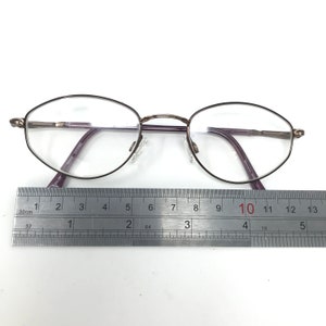Puede incluir: Gafas de forma ovalada con montura oscura y lentes transparentes. Las gafas miden aproximadamente 13 cm de ancho, medidos con una regla. Las patillas son de color morado oscuro. El texto en la patilla dice "TK018 7725507".