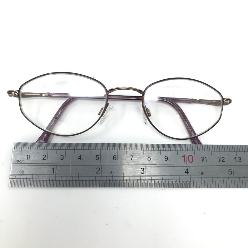 Puede incluir: Gafas de forma ovalada con montura oscura y lentes transparentes. Las gafas miden aproximadamente 13 cm de ancho, medidos con una regla. Las patillas son de color morado oscuro. El texto en la patilla dice "TK018 7725507".