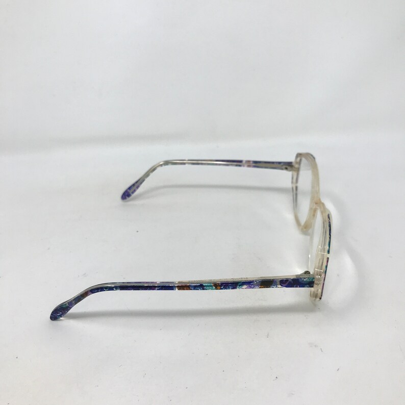 Puede incluir: Gafas con montura transparente y patillas estampadas. Las patillas presentan un dise&ntilde;o colorido con detalles en azul, morado y verde. Las gafas tienen forma de lente redonda y est&aacute;n sobre un fondo blanco.