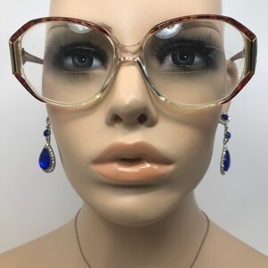 May include: Clear eyeglasses with a light brown and gold frame. The glasses have a large, rounded shape. Dangling blue teardrop earrings with silver accents are visible on either side of the glasses.