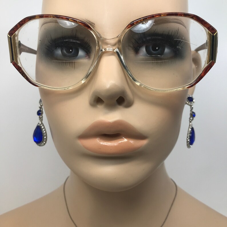 May include: Clear eyeglasses with a light brown and gold frame. The glasses have a large, rounded shape. Dangling blue teardrop earrings with silver accents are visible on either side of the glasses.