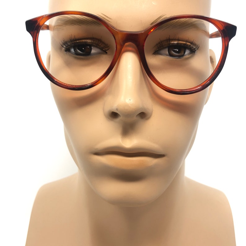 May include: Round-framed eyeglasses with a tortoiseshell pattern. The glasses have a classic design with a dark brown rim and a clear lens. The frames are set against a neutral background.