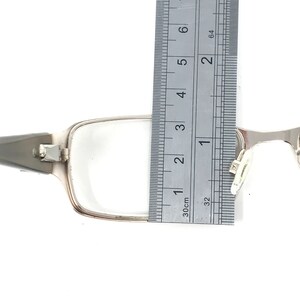 May include: Silver-framed eyeglasses with rectangular lenses. A ruler is placed next to the frame, measuring approximately 1.5 inches in width. The glasses have a hinge and nose pads.