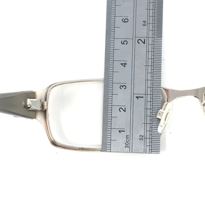 May include: Silver-framed eyeglasses with rectangular lenses. A ruler is placed next to the frame, measuring approximately 1.5 inches in width. The glasses have a hinge and nose pads.