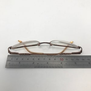 May include: A pair of eyeglasses with a brown metal frame and clear lenses. The glasses are measured with a ruler, showing a length of approximately 5.5 inches. The frame has a classic design.