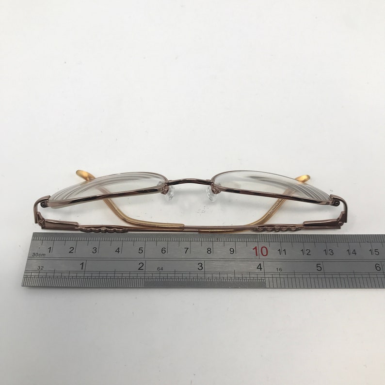 May include: A pair of eyeglasses with a brown metal frame and clear lenses. The glasses are measured with a ruler, showing a length of approximately 5.5 inches. The frame has a classic design.