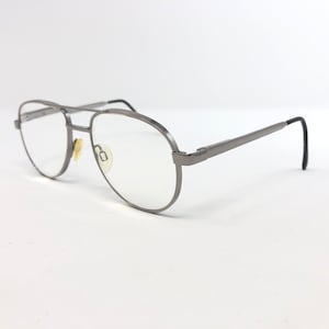 Peut inclure: Lunettes de vue aviateur argent&eacute;es avec verres transparents. Les montures ont un double pont et des branches droites avec des embouts noirs. Les plaquettes nasales sont de couleur jaune clair. Les lunettes sont pos&eacute;es sur fond blanc.