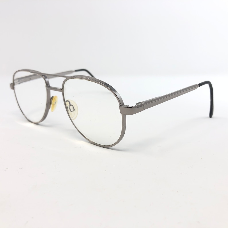 Peut inclure: Lunettes de vue aviateur argent&eacute;es avec verres transparents. Les montures ont un double pont et des branches droites avec des embouts noirs. Les plaquettes nasales sont de couleur jaune clair. Les lunettes sont pos&eacute;es sur fond blanc.