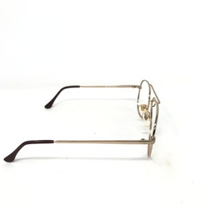 May include: A pair of eyeglasses with a silver metal frame and dark brown temples. The lenses are clear, and the glasses are set against a white background. The glasses are viewed from the side.