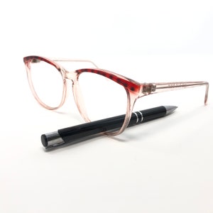 May include: Clear-framed eyeglasses with red tortoise shell accents on the upper rims. A black pen with silver accents rests beneath the glasses. The glasses have a rectangular shape.